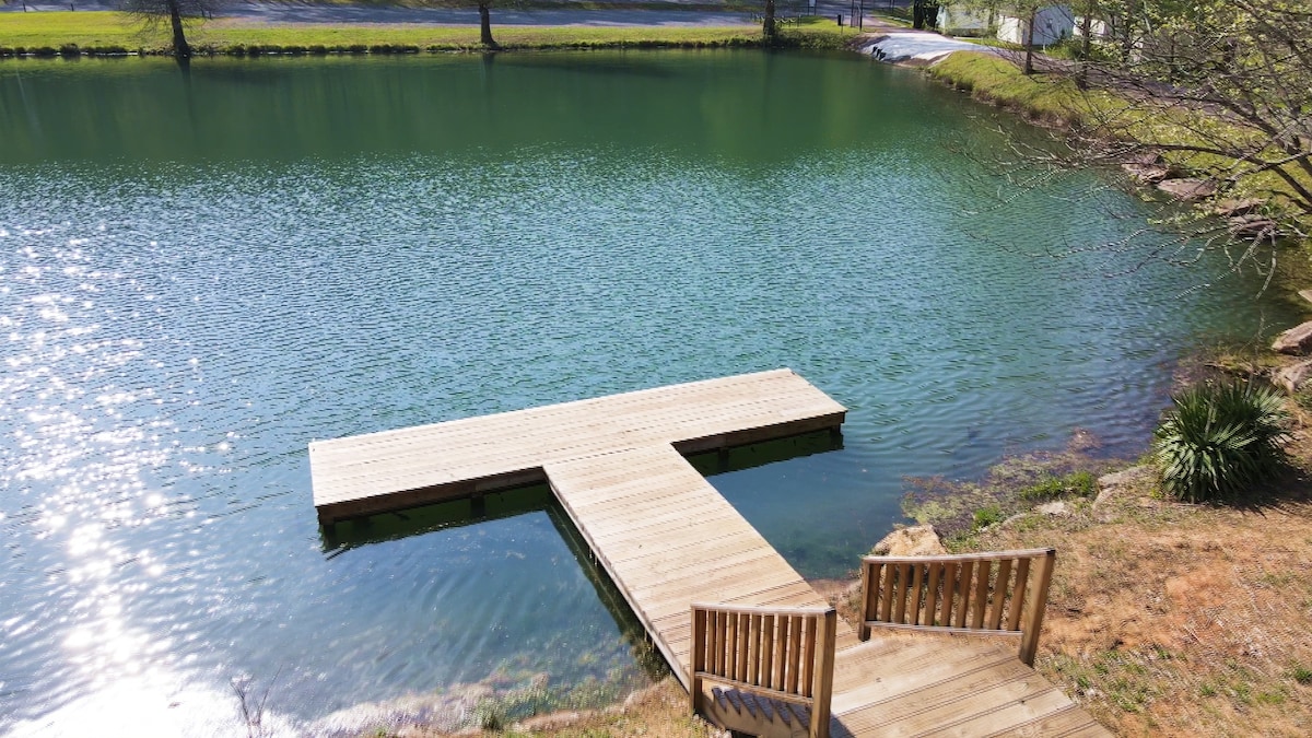 A wooden dock extends into a serene lake, surrounded by lush greenery. Two comfortable chairs are positioned at the edge of the dock, offering a peaceful spot for relaxation. The water reflects sunlight, creating a calming ambiance.