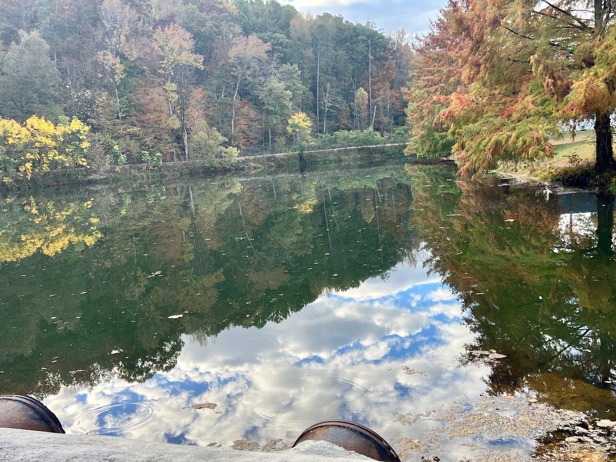 A calm, reflective surface of water is framed by lush trees in autumn colors. The sky is spotted with clouds, creating a serene atmosphere. The tranquil setting invites relaxation and contemplation, perfect for a nature-filled getaway.