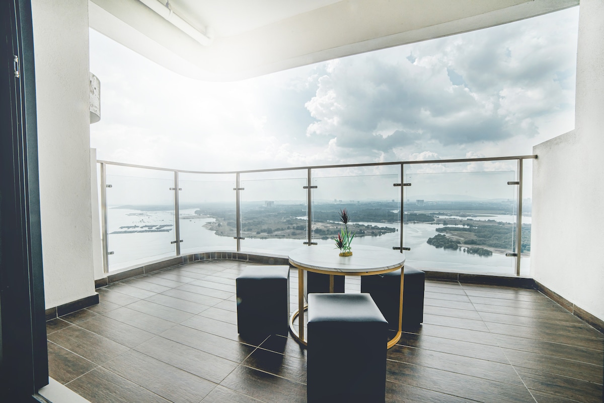 A balcony space offers a full view of the sea, with a table and two square stools positioned on a tiled floor. The railing is made of glass, allowing for unobstructed sightlines of the landscape, which includes water and greenery in the distance.