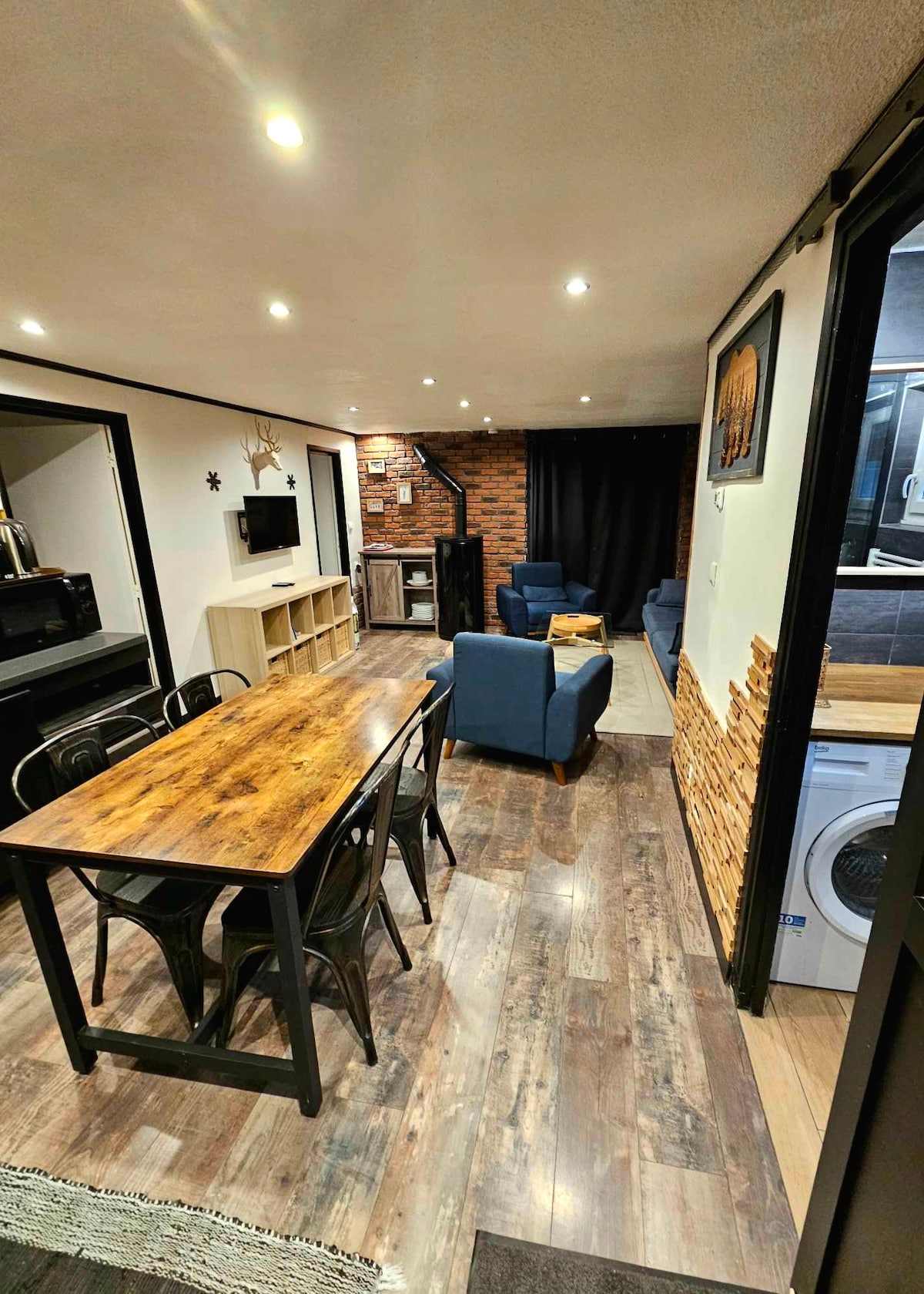 A spacious living area is highlighted by a rustic wooden dining table surrounded by metal chairs. Comfortable blue armchairs are positioned nearby. The decor includes a mix of stone and wood elements, and recessed lighting enhances the ambiance.