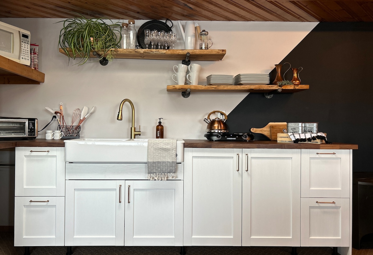 A modern kitchen area is featured with a white farmhouse sink and wooden countertops. Open shelves display dishware and glasses, while kitchen appliances, including a microwave and kettle, are neatly arranged. Minimalist decor creates a clean, functional space.