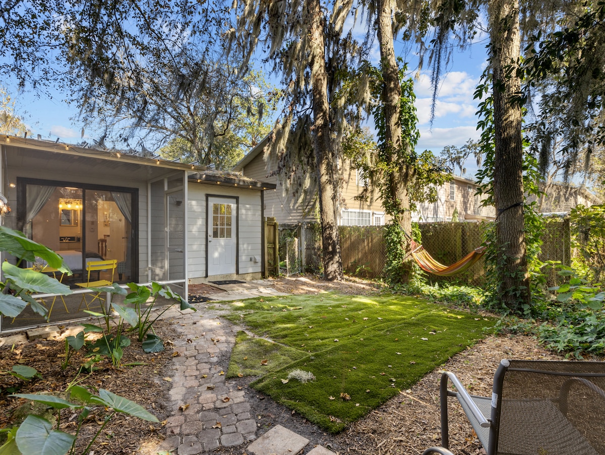 The outdoor space is surrounded by lush greenery and mature trees, providing a serene atmosphere. A small patio area can be seen, featuring a few chairs and a hammock suspended between trees. Well-maintained artificial grass creates a neat space for relaxation or play.