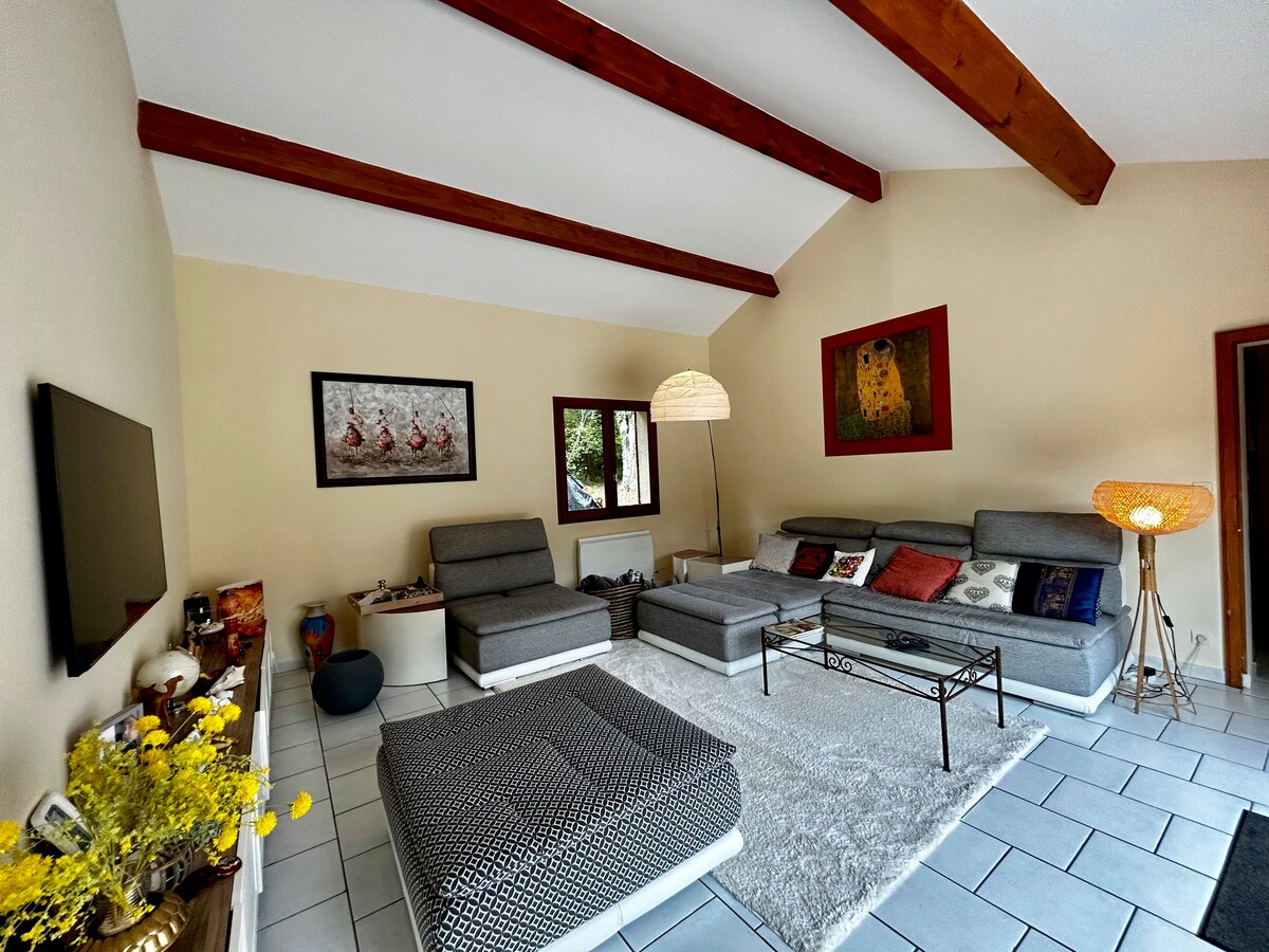 A spacious living area is presented with high, wooden beam ceilings. Comfortable seating arrangements are visible, including a sectional sofa and a lounge chair. A light-colored area rug covers the tiled floor, complemented by decorative wall art and soft lighting from a standing lamp.