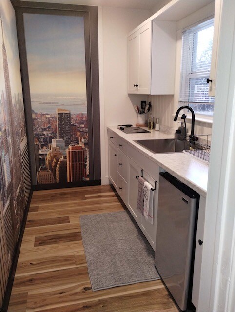 A compact kitchenette features white cabinetry with modern hardware and a sleek countertop. An urban skyline mural adorns the wall, while stainless steel appliances, including a refrigerator, create a contemporary feel. A gray mat lies on the wooden flooring near the sink area.