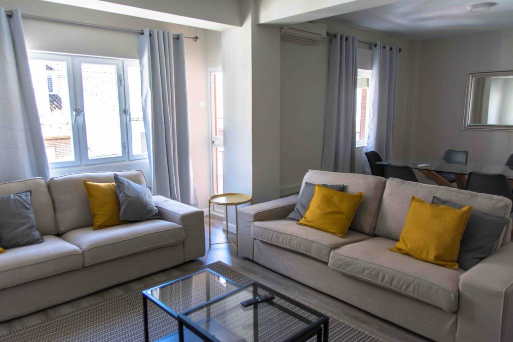 The living area features two light-colored sofas adorned with yellow and gray cushions, positioned around a glass coffee table. Natural light enters through multiple windows, complemented by light curtains. A dining table is visible in the background, accompanied by modern chairs.