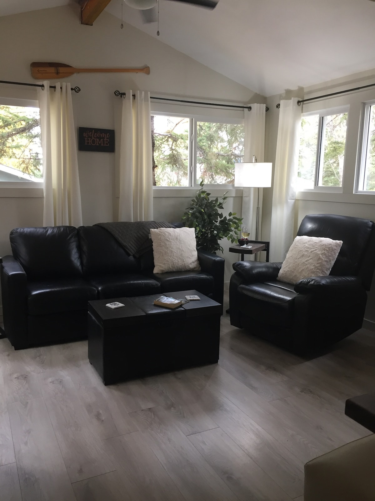 A cozy living area features a black leather sofa and a matching armchair, complemented by decorative pillows. Natural light filters through large windows, with sheer curtains adding a soft touch. A small side table with a lamp and a potted plant enhance the welcoming ambiance.
