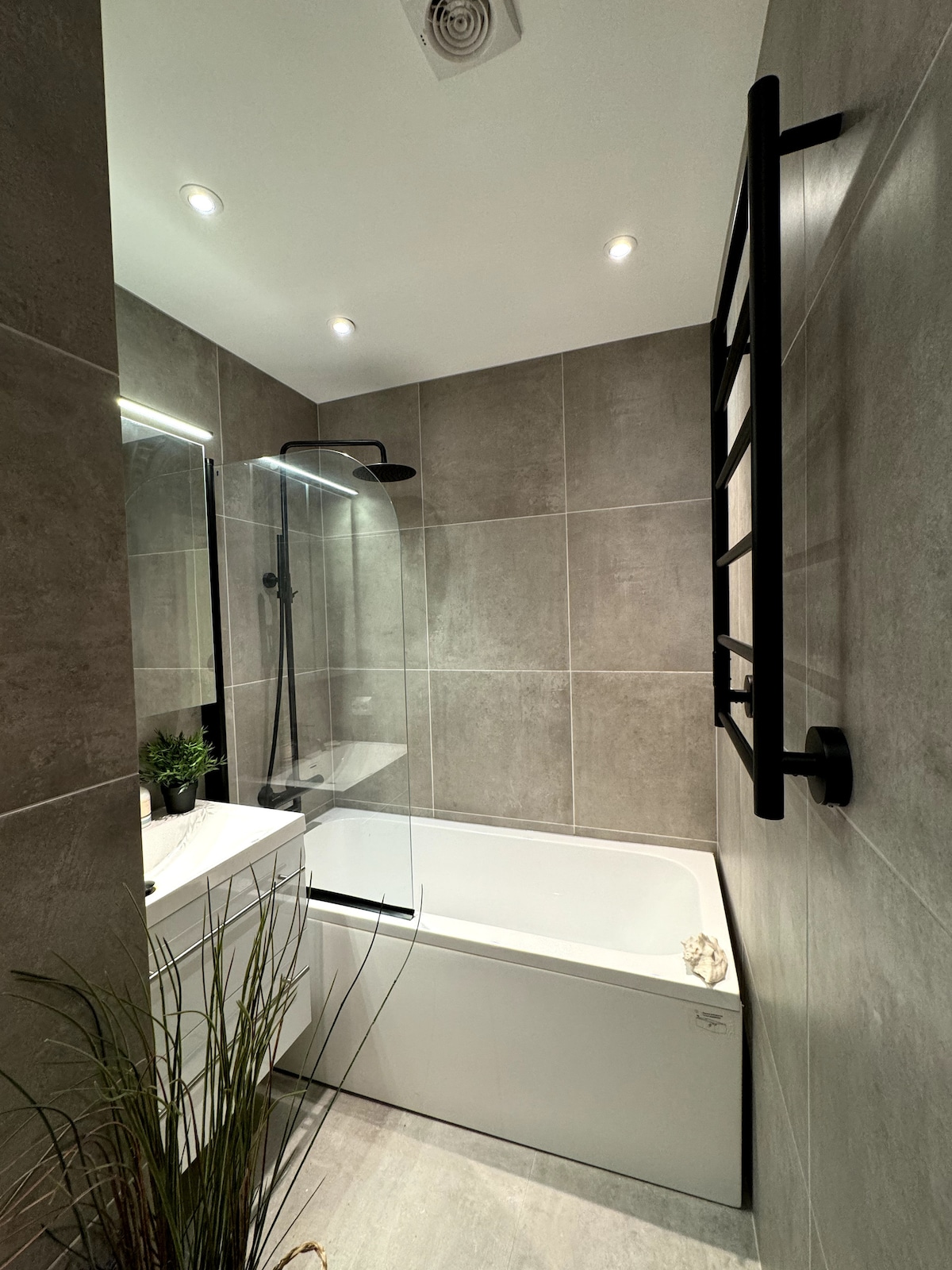 A modern bathroom features gray tiled walls and a glass shower enclosure. A white bathtub is positioned beside a compact sink with a mirror above. Soft lighting is provided by recessed ceiling fixtures, and a decorative plant adds a touch of nature.