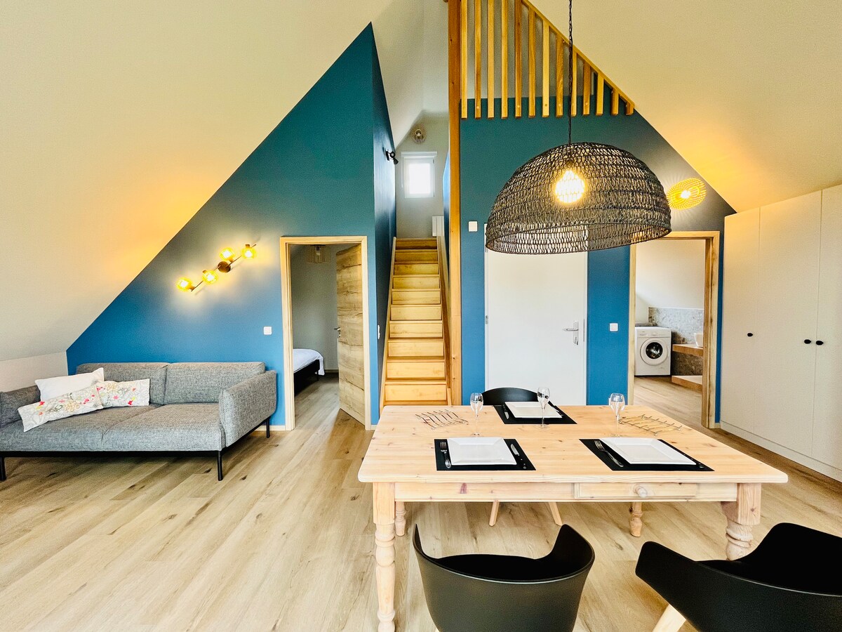 An airy interior is displayed with a light wood dining table set for two, accompanied by black chairs. A cozy gray sofa sits to the left, while warm light fixtures illuminate the blue walls. A staircase leads to the upper level, enhancing the open layout.