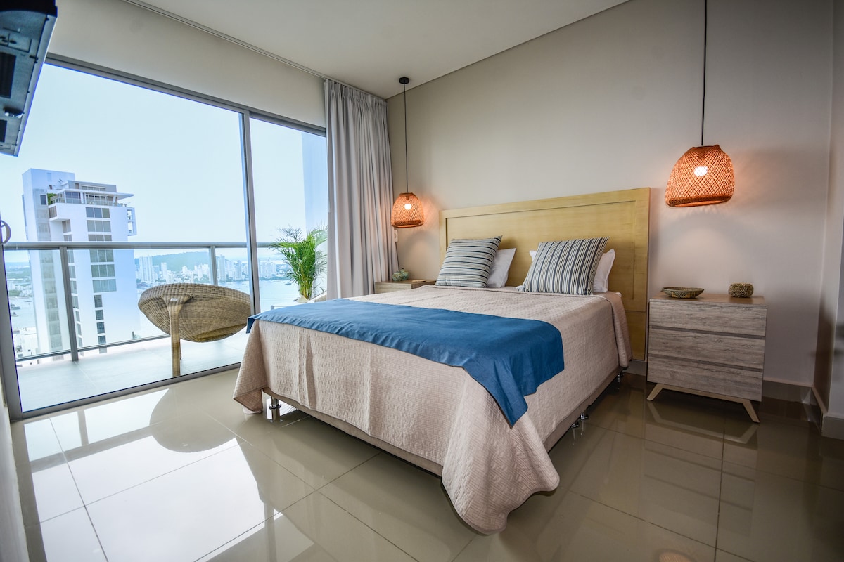 A spacious bedroom is highlighted by large windows offering panoramic views of the city and bay. A Queen-sized bed is adorned with soft linens, complemented by two pendant lights. A seating area with a woven chair adds to the room's comfort and elegance.