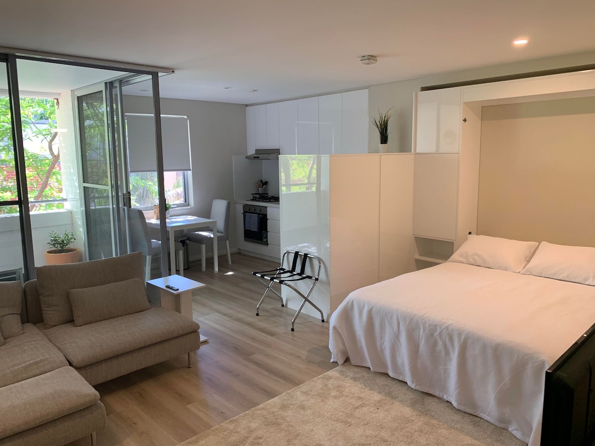 An open-plan living area features a light-colored sofa facing a spacious balcony. A dining table is set against the wall, complemented by a fully equipped kitchen with modern appliances. A cozy sleeping area includes a spacious bed, visually separated by a white partition.