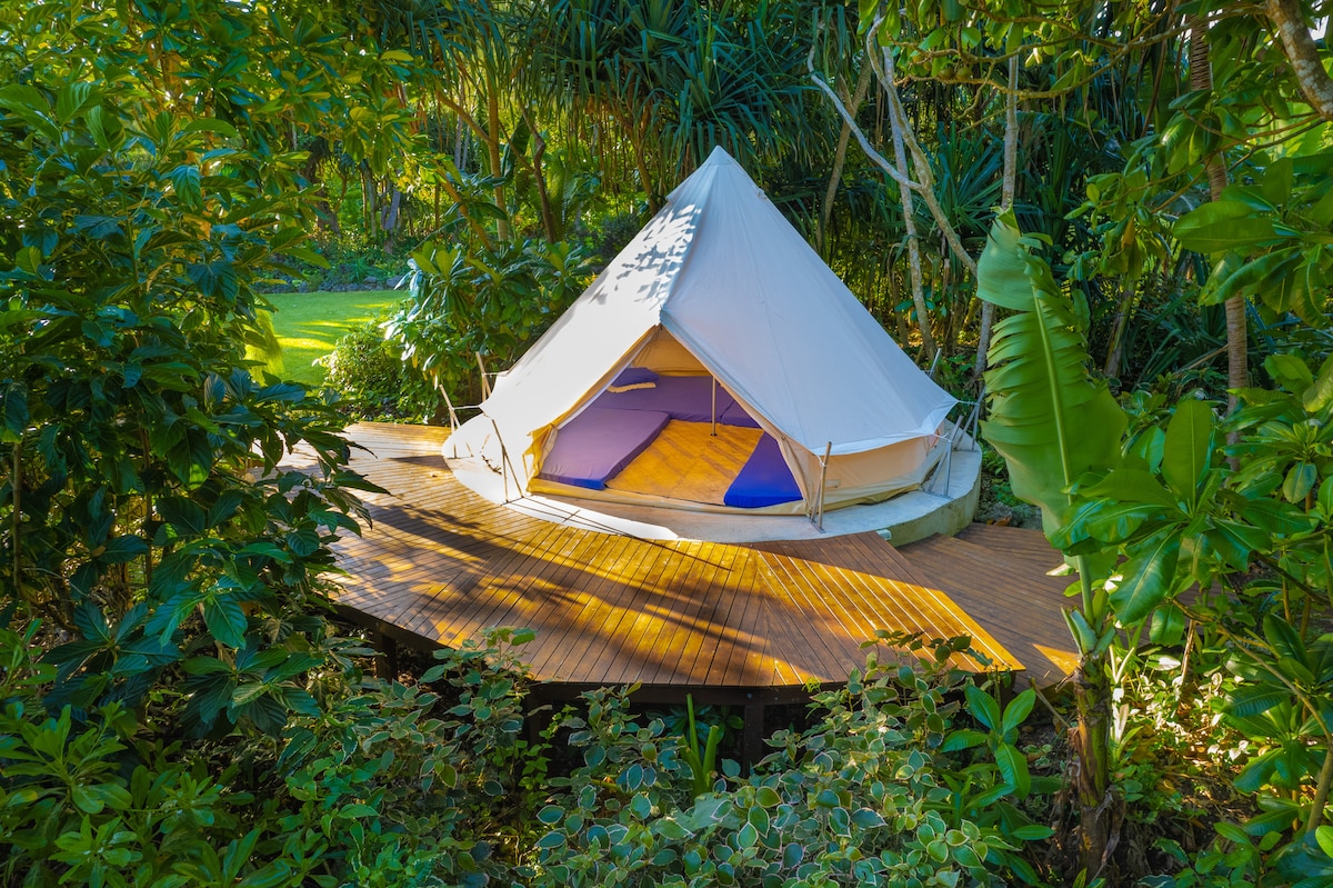 A spacious glamping tent is set on a wooden platform surrounded by lush greenery. The tent, featuring a conical shape, is partially opened to reveal comfortable futons inside. Natural light filters through the leaves, creating a serene atmosphere around the site.