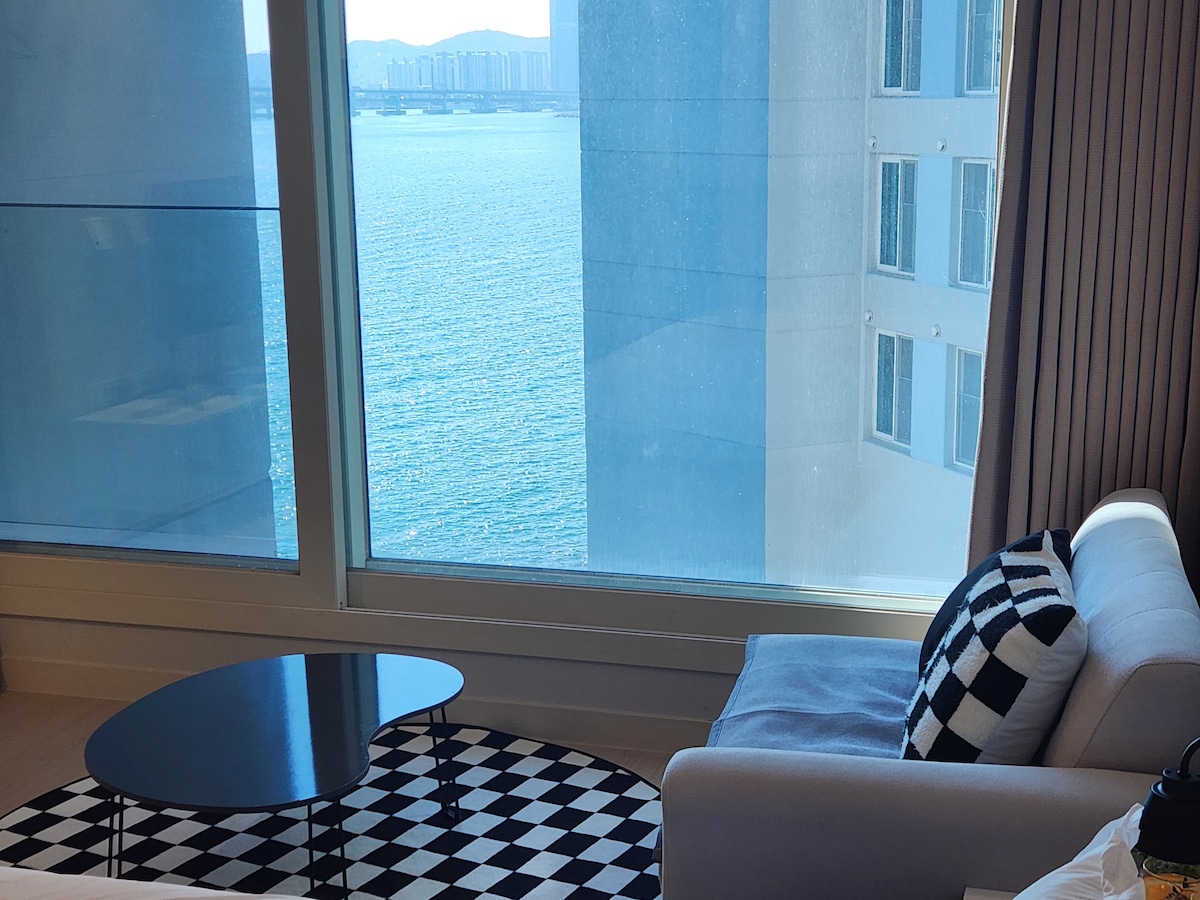 A contemporary seating area is positioned by a large window, offering views of the ocean. A sleek, round coffee table is centered on a monochrome checkered rug, while a light-colored sofa adorned with striped cushions invites relaxation. Natural light fills the space, enhancing its airy feel.