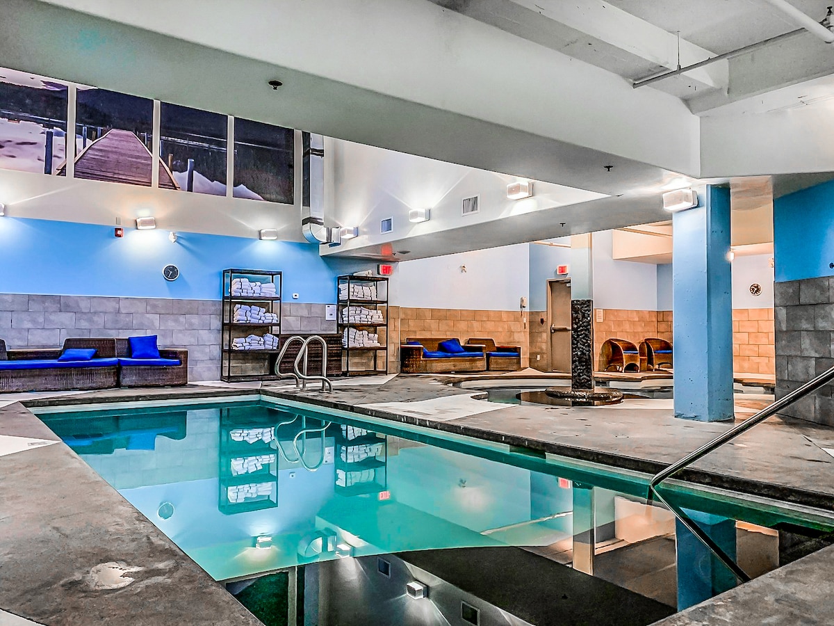 An indoor pool area features a rectangular pool with clear water. Surrounding the pool, seating areas with blue cushions are provided. Soft lighting reflects off the water's surface, and a spa-like hot tub can be seen in the corner, contributing to a calm ambiance.