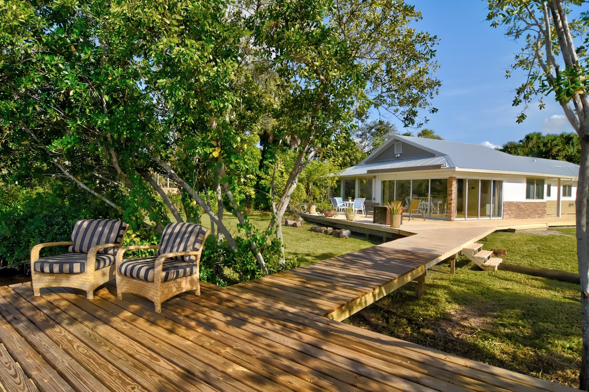 A wooden deck extends toward a well-maintained home surrounded by lush greenery. Two striped chairs are positioned on the deck, offering a place to relax and enjoy the scenic views of the natural setting.