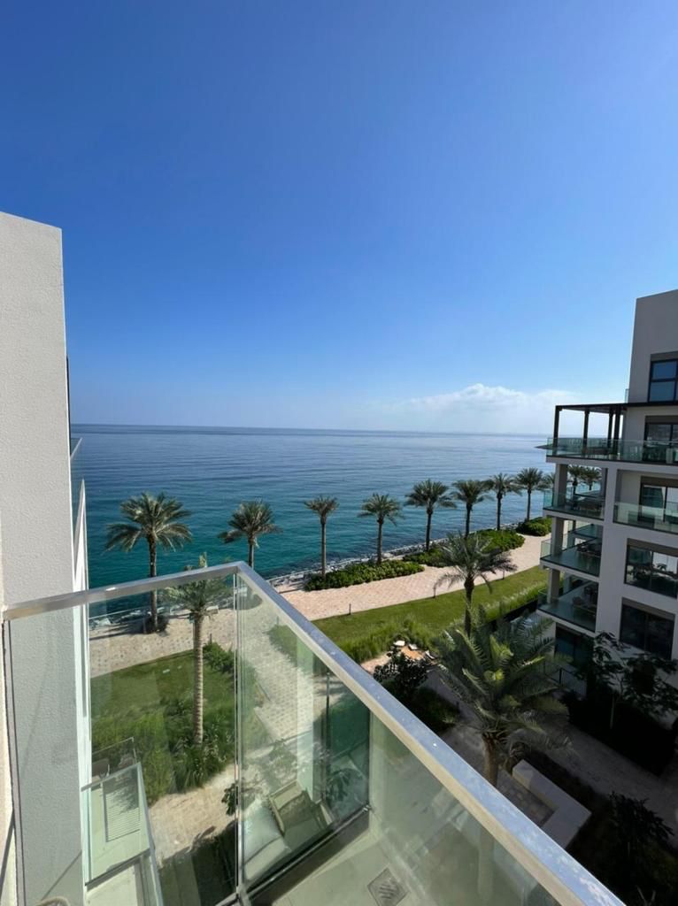 A balcony offers a clear view of the coastline, featuring a calm sea meeting a bright blue sky. Palm trees are visible along the sandy beach, complemented by manicured gardens in the foreground, creating a serene coastal atmosphere.