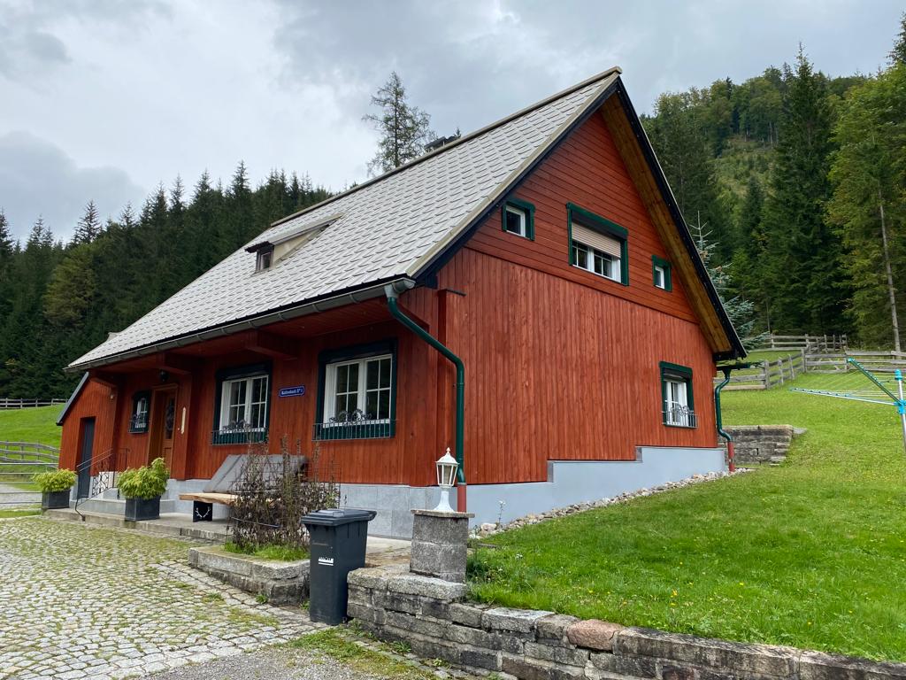 A charming chalet is shown featuring a warm wooden exterior. Grass surrounds the structure, and trees are present in the backdrop. The roof has a sloping design with a small dormer visible, and light streams through the windows. An outdoor pathway leads to the entrance.