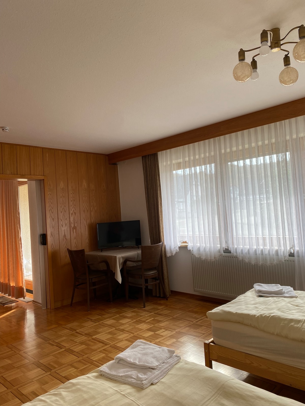 A spacious room features wooden accents and large windows with sheer curtains allowing natural light. A small dining table is set with two chairs, and a television is positioned nearby. Fresh linens are neatly arranged on the beds, contributing to a clean and welcoming environment.