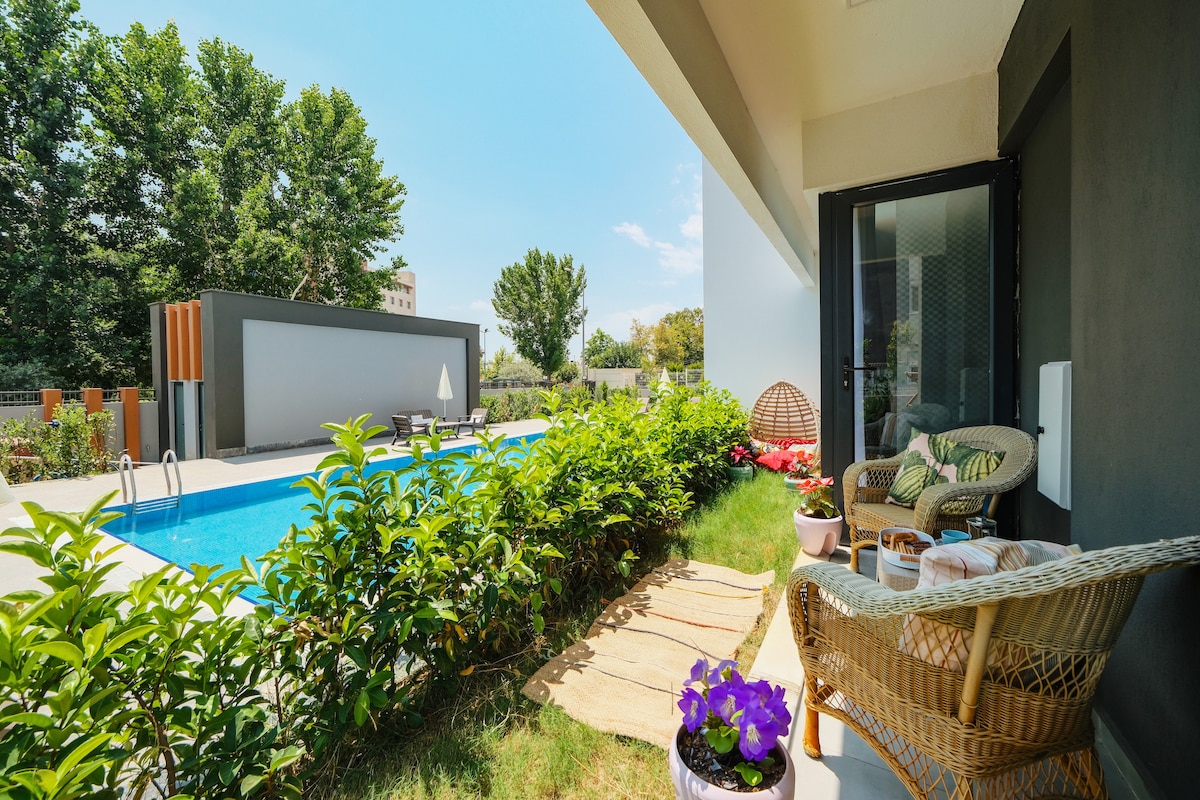 A serene patio area is adjacent to the pool, surrounded by lush greenery. Comfortable wicker seating allows for relaxation. Colorful potted flowers add a touch of nature, while a soft rug enhances the welcoming atmosphere of the space.