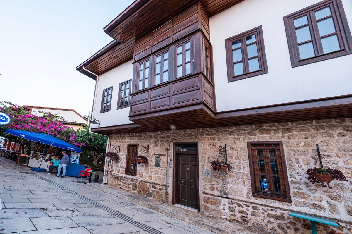 An elegant building features a mix of stone and wood elements, showcasing traditional architecture. Large windows adorned with wooden frames provide a glimpse into the structure. Decorative flower boxes enhance the exterior, while a cobblestone pathway leads to nearby outdoor seating areas.
