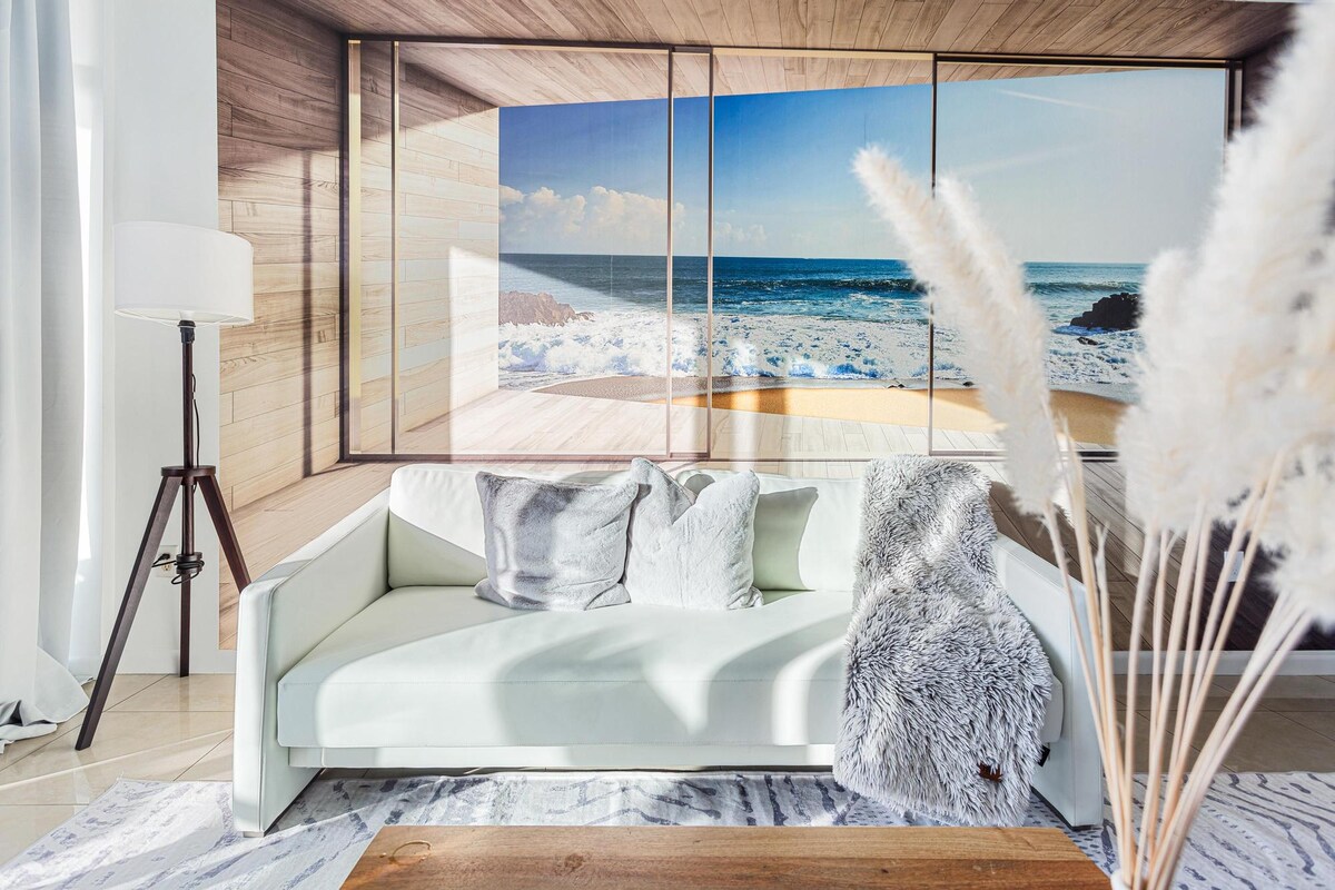 A modern living area is depicted, featuring a sleek white sofa adorned with pillows and a soft throw blanket. A large window provides a view of the ocean, while natural light illuminates the space. A floor lamp stands beside the sofa, enhancing the bright atmosphere.