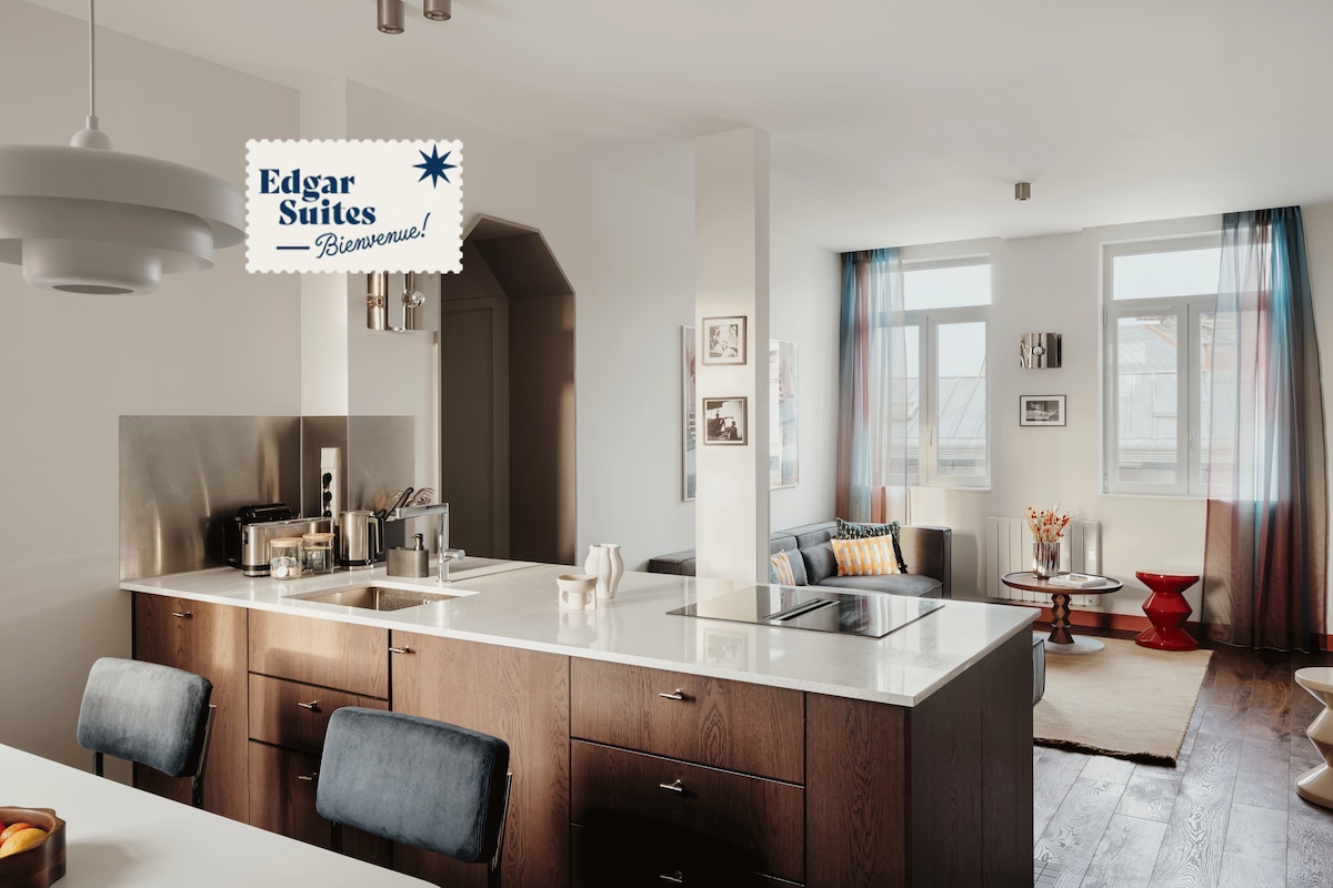 A modern kitchen features a sleek island with a white countertop, equipped with a stovetop and stylish bar stools. Natural light floods the space through large windows, creating an airy atmosphere. A cozy seating area with a sofa and coffee table is visible in the background.