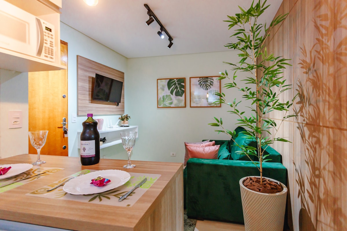 A cozy dining area is set with decorative plates and glasses. A leafy plant adds a touch of greenery. A comfortable sofa with cushions is visible, alongside wall art depicting tropical leaves. Natural light illuminates the space, enhancing the inviting ambiance.