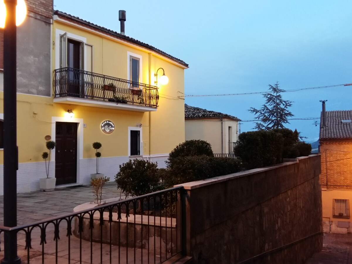 A charming yellow building is featured, with a balcony adorned with potted plants. Soft evening light illuminates the exterior. Surrounding greenery and a low stone wall enhance the entrance, while a walkway leads up to the door, creating an inviting atmosphere.