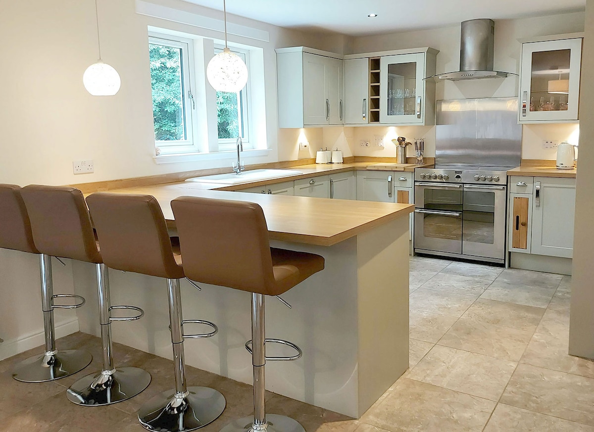 A modern kitchen is highlighted by sleek cabinetry and stainless steel appliances. A central island with seating for four is complemented by stylish pendant lighting. Large windows allow natural light to illuminate the space, featuring a neutral tile floor that enhances the clean design.