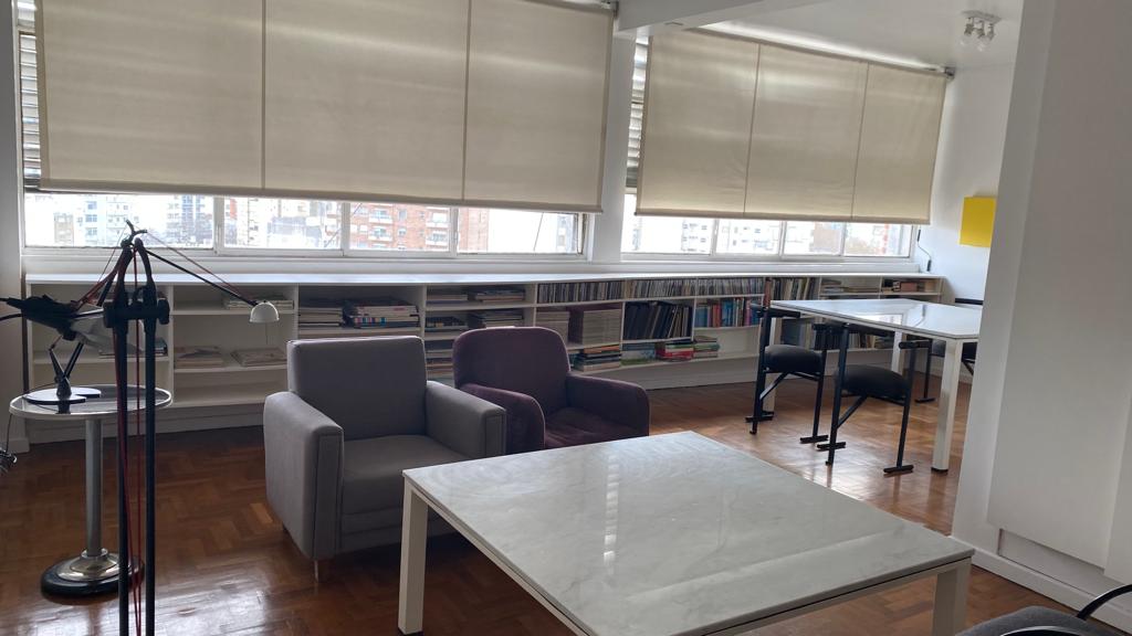 A well-lit living area features a white marble table positioned between two armchairs. Shelves filled with books line the wall behind, while natural light floods in through large windows, covered by soft beige shades.