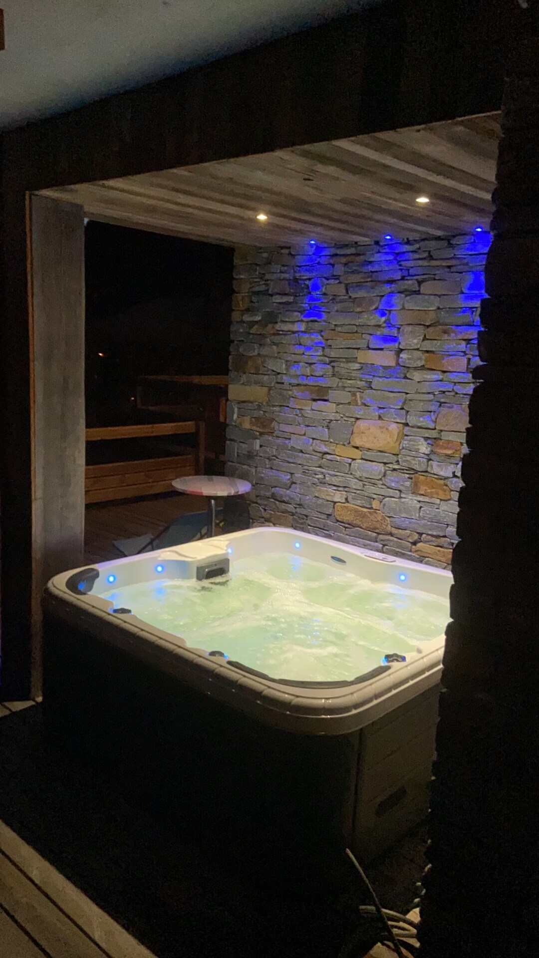 A private jacuzzi is situated against a stone wall, illuminated by blue lights. The soothing water reflects the soft glow, creating a serene ambiance. This space is partially enclosed, providing an inviting atmosphere for relaxation and enjoyment.