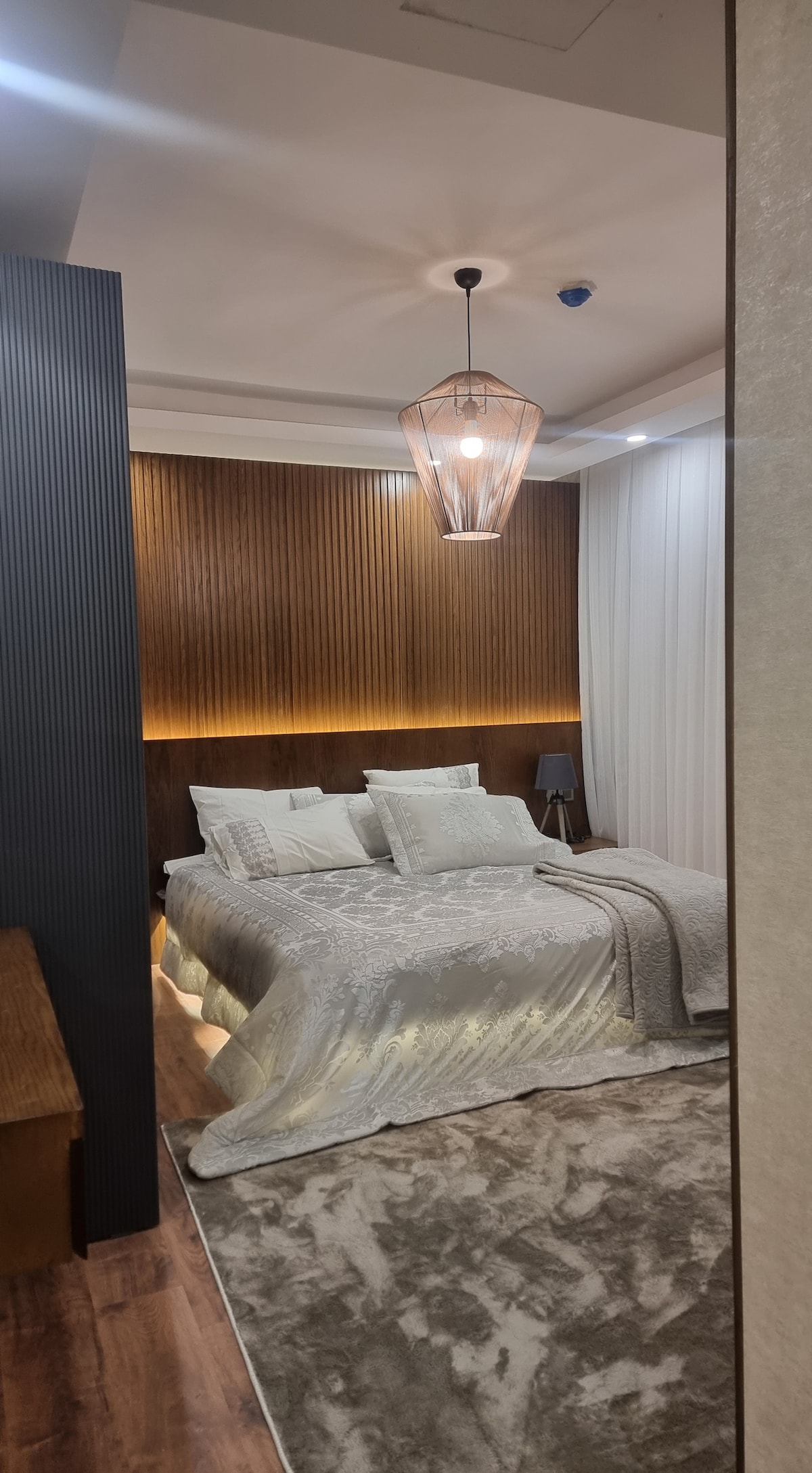 A serene bedroom is presented, featuring a spacious bed adorned with textured bedding and decorative pillows. Soft lighting emanates from a modern pendant fixture above, contrasting with the warm wooden paneling. A plush area rug complements the room's cozy atmosphere.