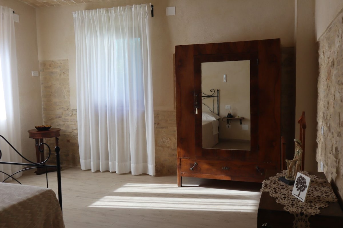 A serene bedroom is featured, showcasing a wooden wardrobe with a mirrored front against a stone wall. Soft, white curtains diffuse natural light, and light-colored flooring complements the space. A glimpse of the bed is visible in the background, enhancing the room's tranquil ambiance.