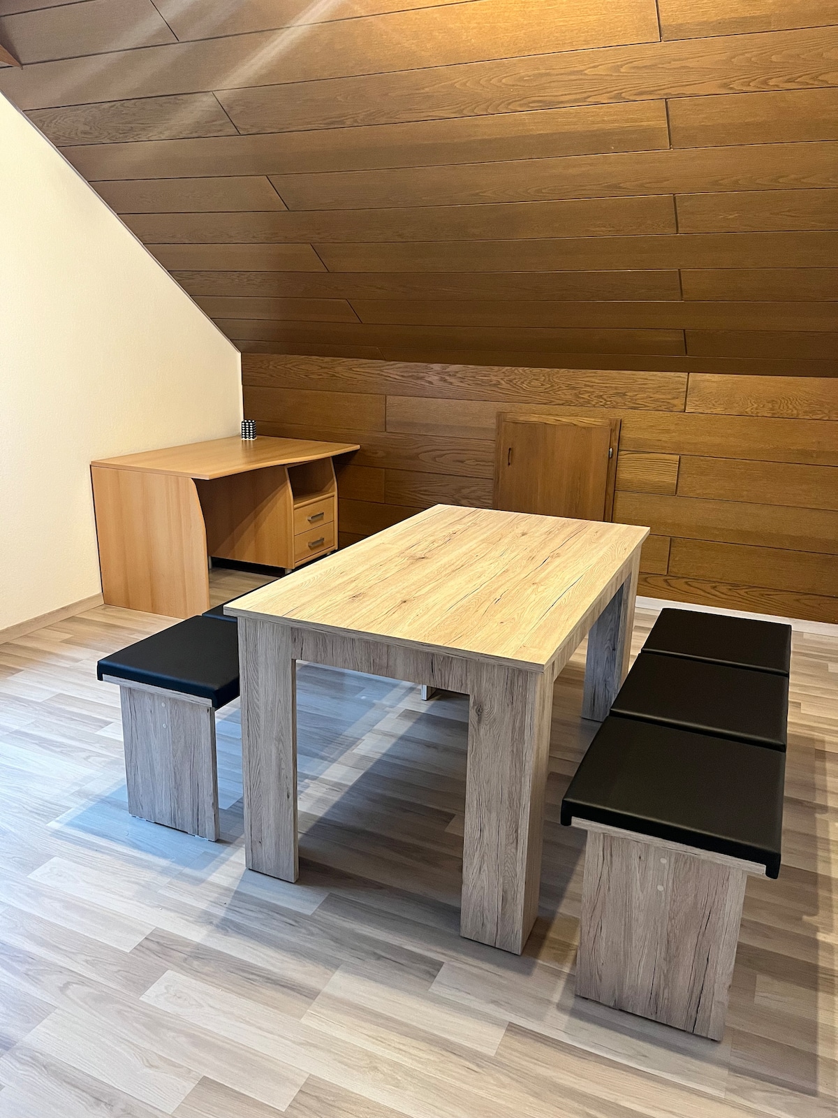 A simple dining area features a wooden table surrounded by black benches. A complementary desk is visible against the wall, with drawers for storage. The sloped wooden ceiling adds a warm touch to the space, which is well-lit with natural light.