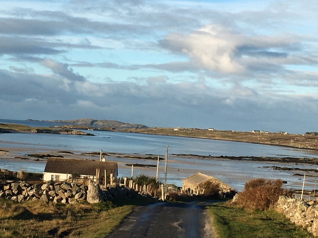 A serene coastal view captures the gentle curve of the shoreline, with small cottages nestled along a winding road. The calm waters reflect a soft blue sky dotted with clouds, while hills rise in the distance, creating a peaceful rural landscape.