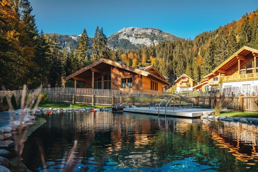 A natural swimming pool is framed by manicured greenery and wooden fencing, reflecting the surrounding mountains and trees. The chalet features a warm wooden exterior, with large windows inviting natural light. The tranquil landscape creates a serene environment, perfect for relaxation.
