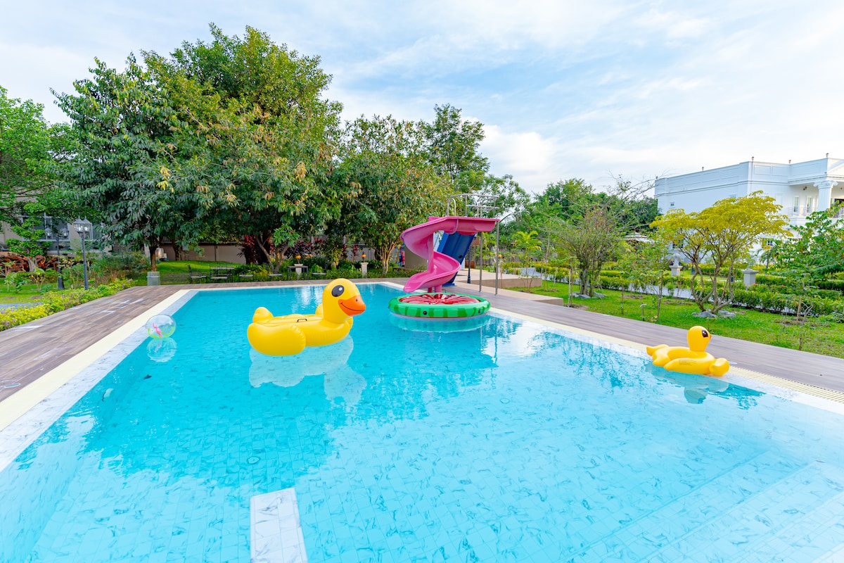 A large swimming pool is seen with a vibrant water slide and several colorful inflatable toys floating on the surface. Lush greenery surrounds the pool, contributing to an inviting outdoor environment.