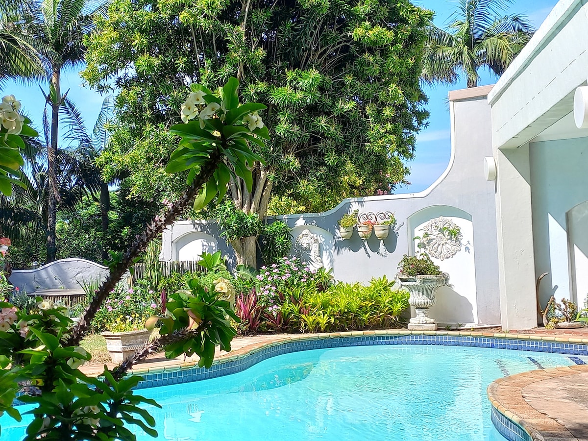 A serene outdoor area is highlighted by a clear blue swimming pool, surrounded by vibrant tropical foliage. A tall tree provides shade, while decorative elements, including wall planters, enhance the garden setting. The pool's gentle curves invite relaxation in a tranquil environment.