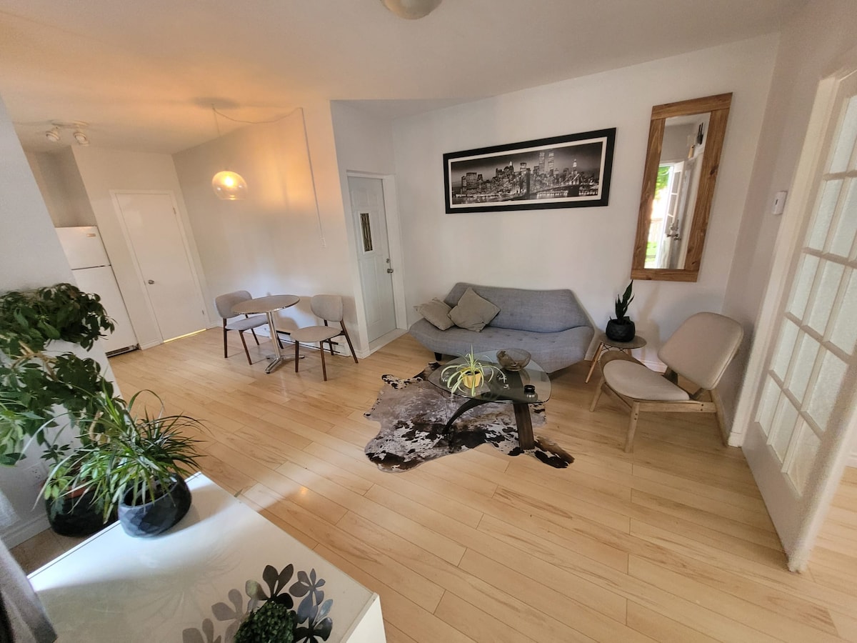 A light-filled living space features a cozy sofa and two comfortable chairs arranged around a glass coffee table atop a patterned area rug. A large mirror and a cityscape artwork accentuate the wall, while plants provide a touch of greenery.