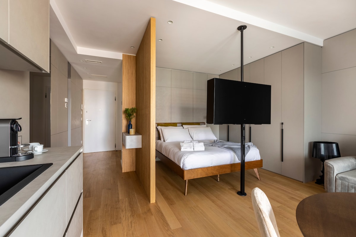 An open space layout features a spacious bed with a wooden frame, a mounted flat-screen TV positioned on a black pole, and sleek cabinetry. Soft natural light fills the area through large windows, creating a bright and airy ambiance complemented by warm wooden flooring.