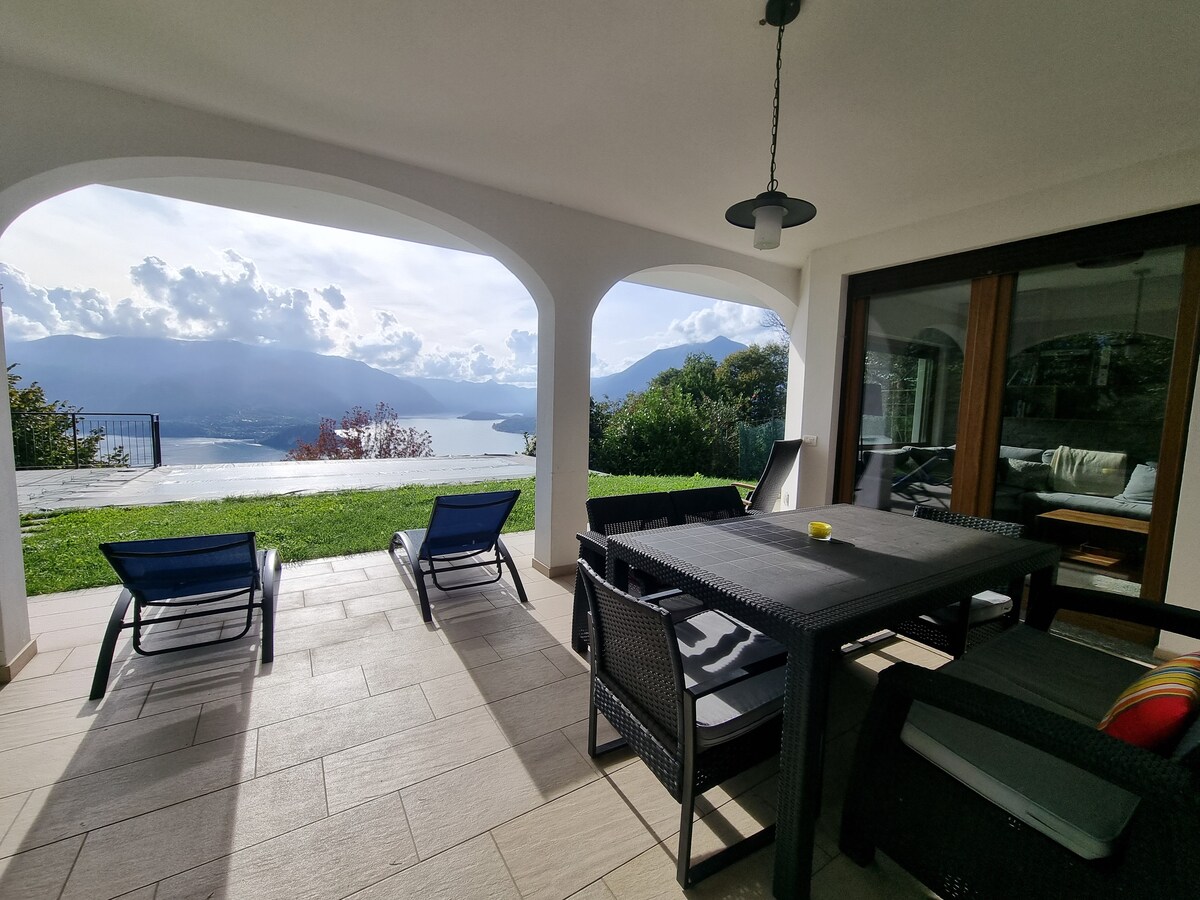 A covered patio features a dining table with seating and two lounge chairs positioned for lake views. Archways frame the scenic landscape of Lake Como, with lush greenery and mountains visible in the background. Natural light brightens the space, enhancing the tranquil atmosphere.