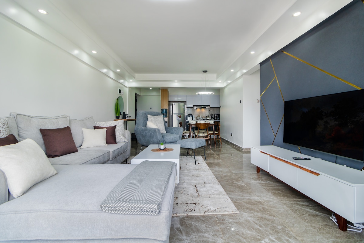 A spacious living area features a large sectional sofa with neutral cushions and a comfortable armchair. A sleek white coffee table is positioned in front of the sectional. The minimalist entertainment unit holds a flat-screen TV, while a dining area with wooden chairs is visible in the background.