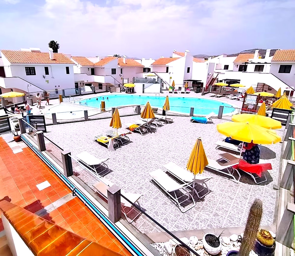 A spacious pool area is surrounded by lounge chairs and yellow umbrellas. The inviting swimming pool stretches across the center, with well-maintained paving leading to surrounding white-washed buildings. A vibrant atmosphere is created by sunny weather and poolside amenities.
