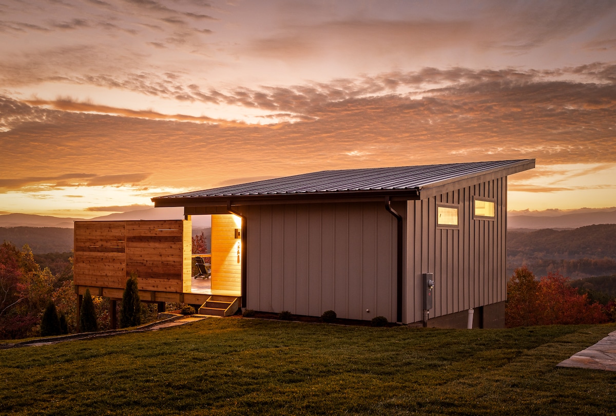 The studio cabin is highlighted against a colorful sunset, featuring a contemporary design with a sloped roof and wooden accents. The outdoor space includes a small deck with seating visible, surrounded by rolling green hills and trees.