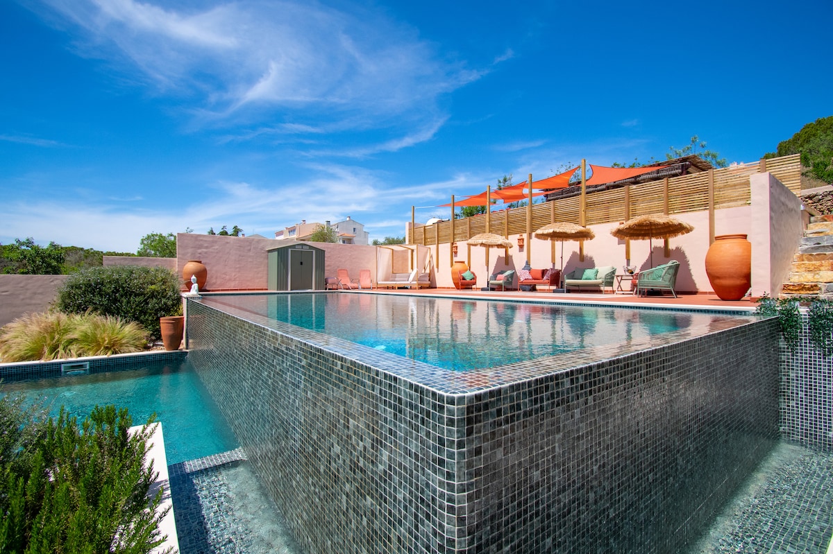 A two-tiered swimming pool is showcased, featuring a stunning infinity edge that reflects the clear blue sky. Surrounding the pool are comfortable lounge chairs and shaded areas with umbrellas, complemented by landscaped greenery and terracotta pots, creating a serene outdoor retreat.