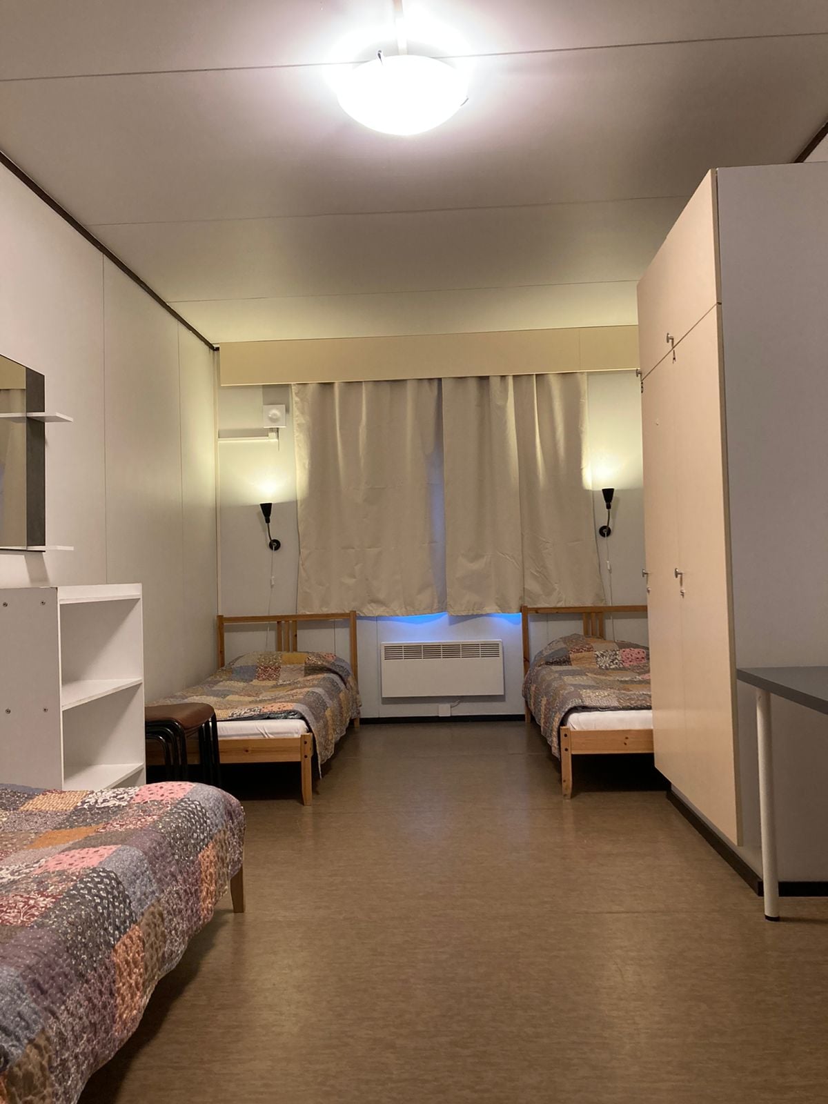 The room is fitted with two beds arranged parallel, each covered with patterned quilts. A large window is dressed with light curtains, allowing soft natural light into the space. A wardrobe and a small table are positioned along the walls, providing functional storage and work areas.