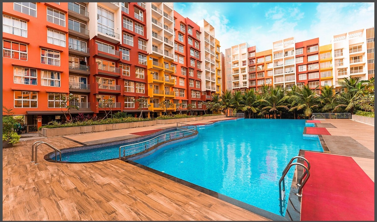 A spacious outdoor area features a large swimming pool framed by vibrant red and orange apartment buildings. Lush palm trees surround the pool deck, which includes comfortable lounging spaces and pathways, creating a relaxed atmosphere for leisure and enjoyment.
