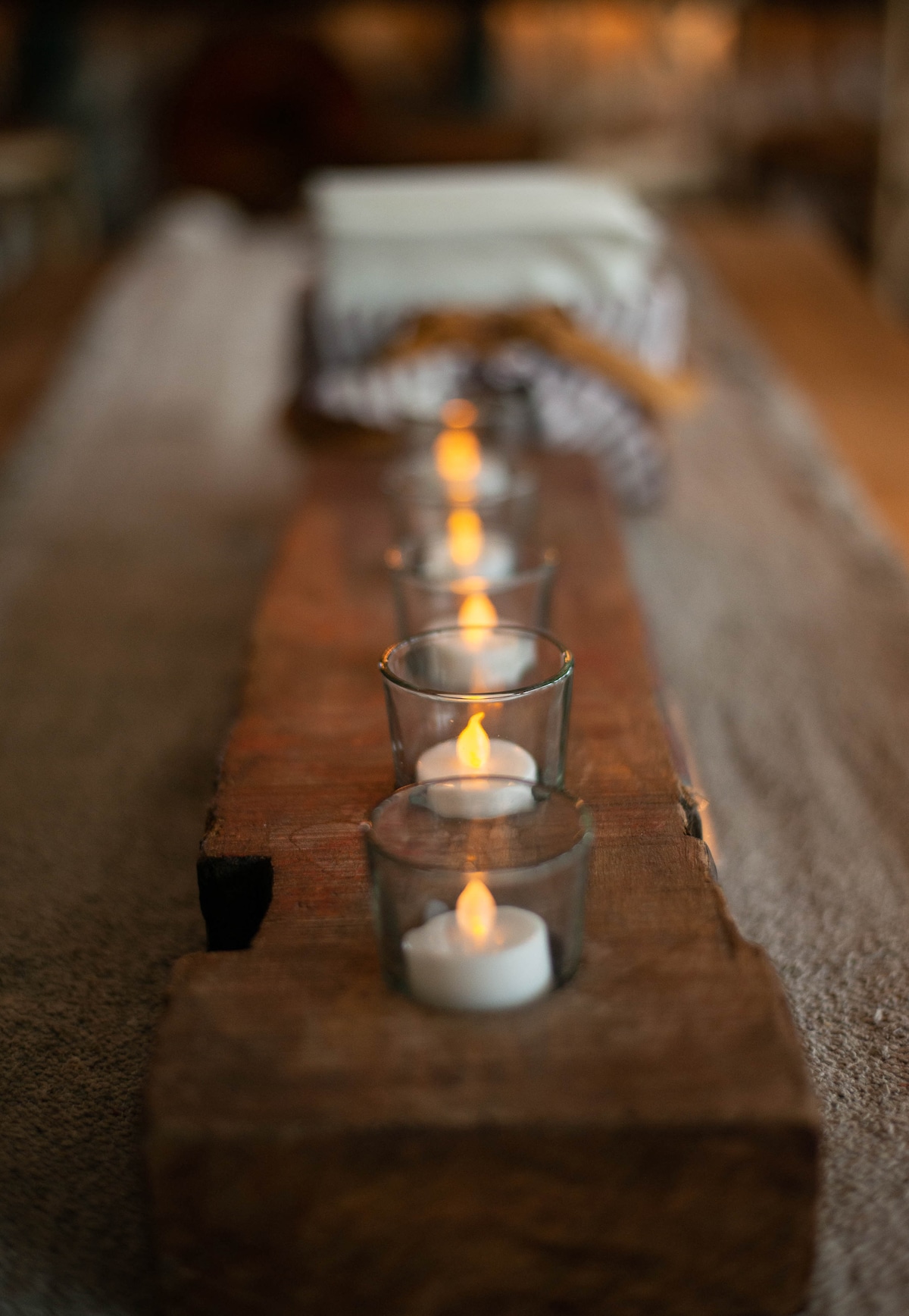 A rustic wooden centerpiece showcases five glass candle holders, each containing a lit candle. The warm glow of the flames is set against a softly textured table runner, creating an inviting focal point for gatherings.