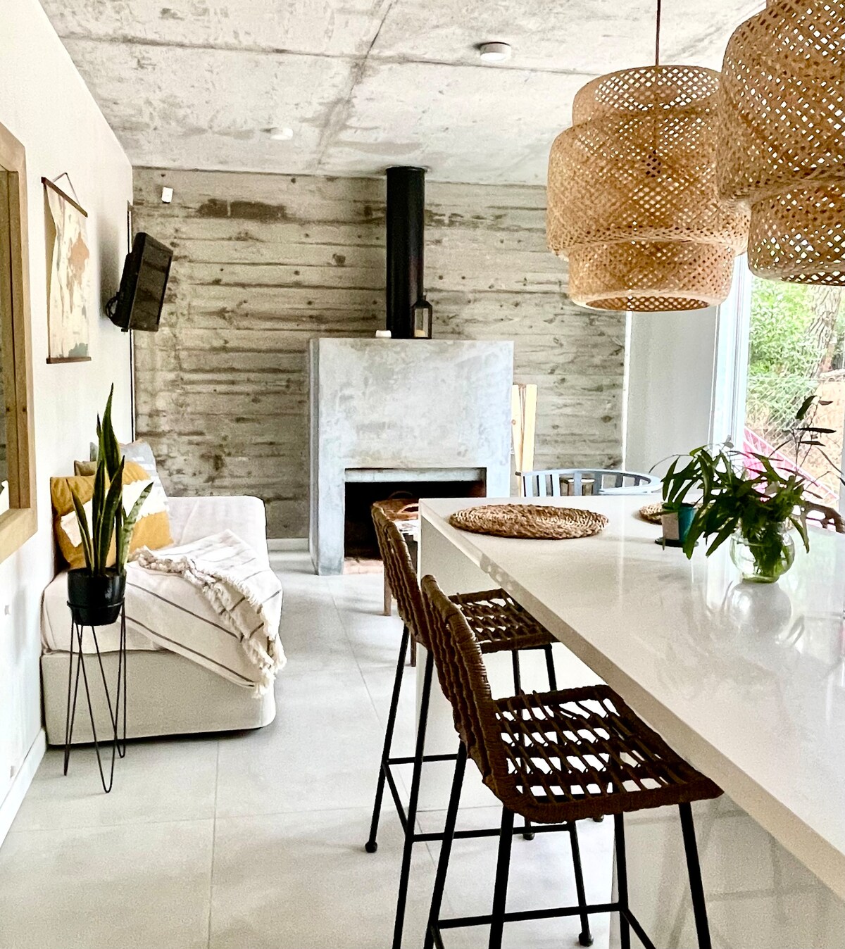 A modern living space features a textured concrete wall and a cozy seating area with a white sofa. A sleek, gray fireplace stands prominently, surrounded by greenery. Natural light floods in through large windows, while woven pendant lights hang above a white kitchen island with bar seating.