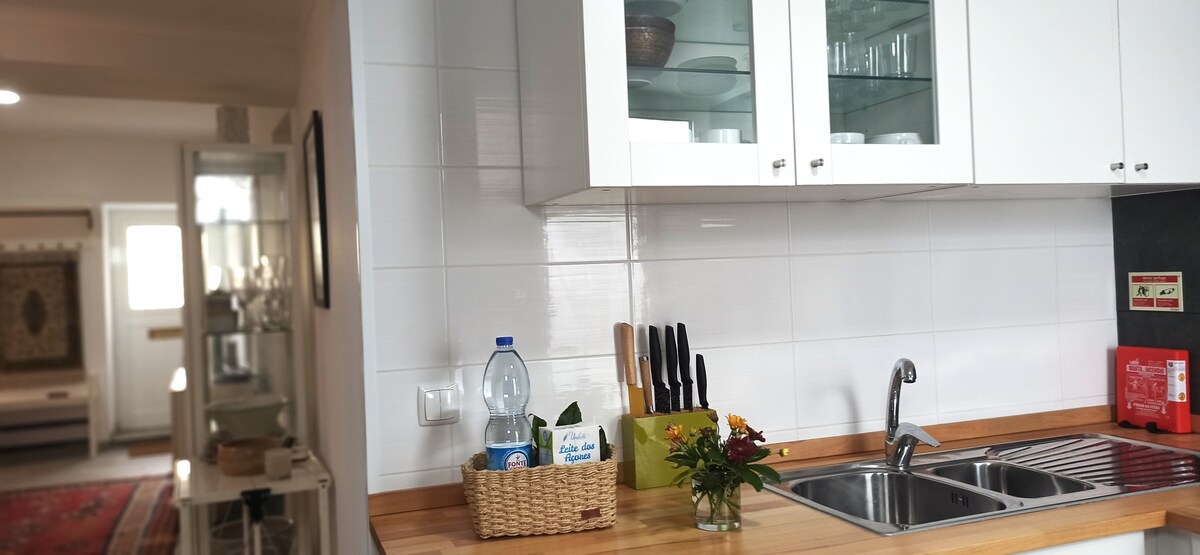 A modern kitchen area is presented, featuring a clean layout with white cabinetry and a wooden countertop. The sink is positioned beside a set of neatly arranged utensils and a matching wicker basket. Glass cabinets display selected kitchenware, contributing to a functional and tidy space.