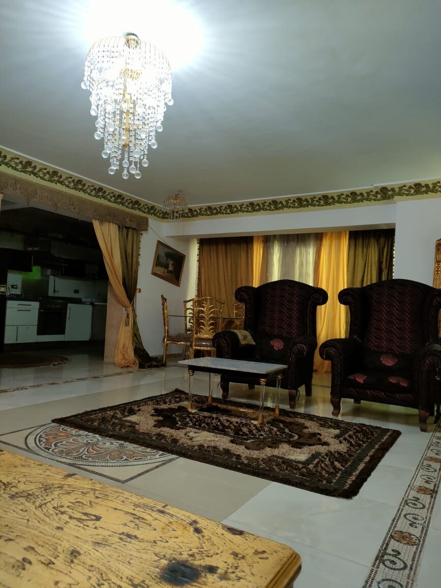 A spacious living area features two ornate chairs with rich upholstery positioned around a central glass table. A decorative rug lies on the polished floor under soft lighting from a crystal chandelier above, complemented by elegant drapes that enhance the overall decor.